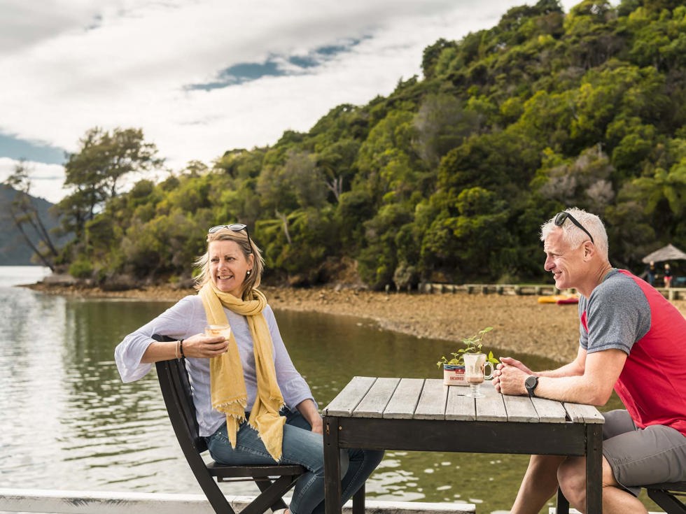 A relaxed couple drink and enjoy Boatshed Cafe and Bar coffee on the jetty at Punga Cove in the Marlborough Sounds in New Zealand