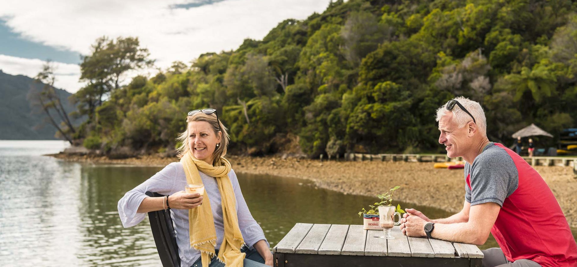 A relaxed couple drink and enjoy Boatshed Cafe and Bar coffee on the jetty at Punga Cove in the Marlborough Sounds in New Zealand
