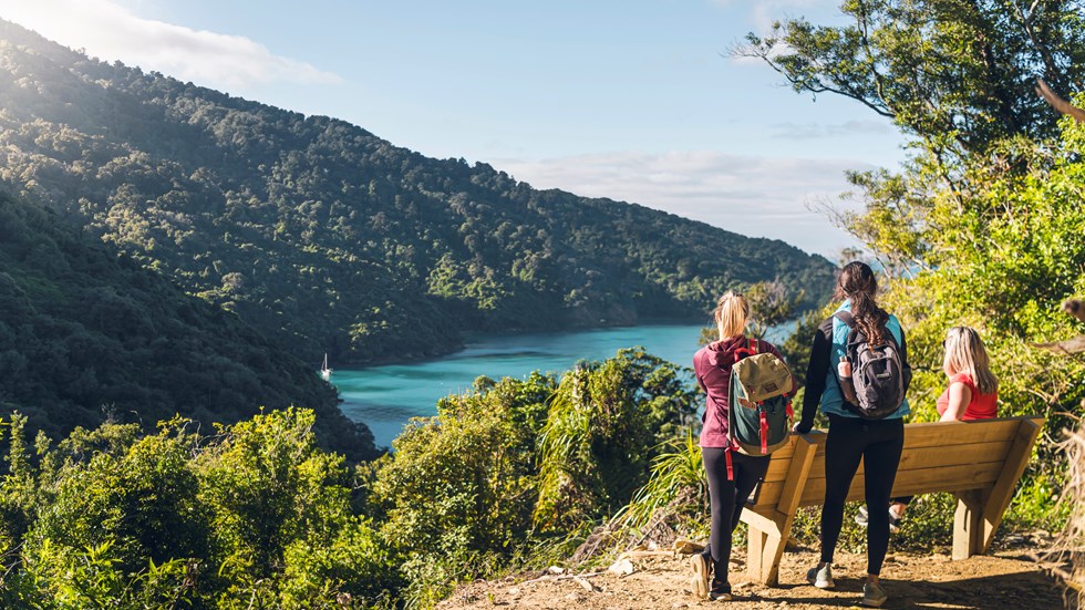 Three people at a bench and viewpoint on the northern Queen Charlotte Track, overlooking Ship Cove/Meretoto in the Marlborough Sounds, New Zealand.