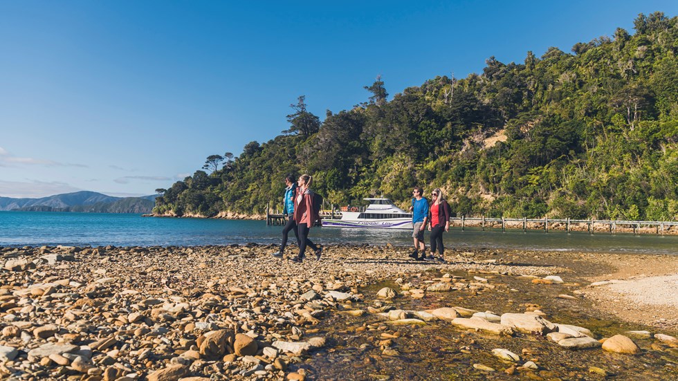 Four people walking on the beach at Ship Cove/Meretoto with a Cougar Line boat in the Marlborough Sounds, New Zealand.