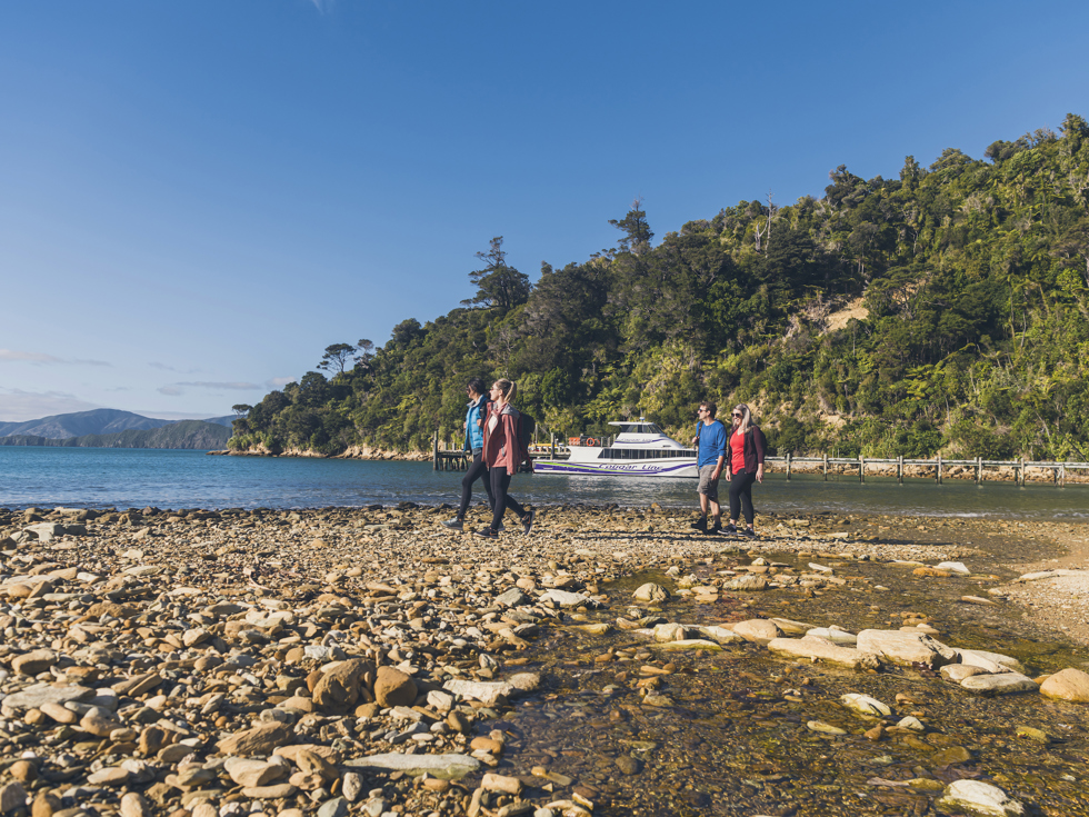 Four people walking on the beach at Ship Cove/Meretoto with a Cougar Line boat in the Marlborough Sounds, New Zealand.