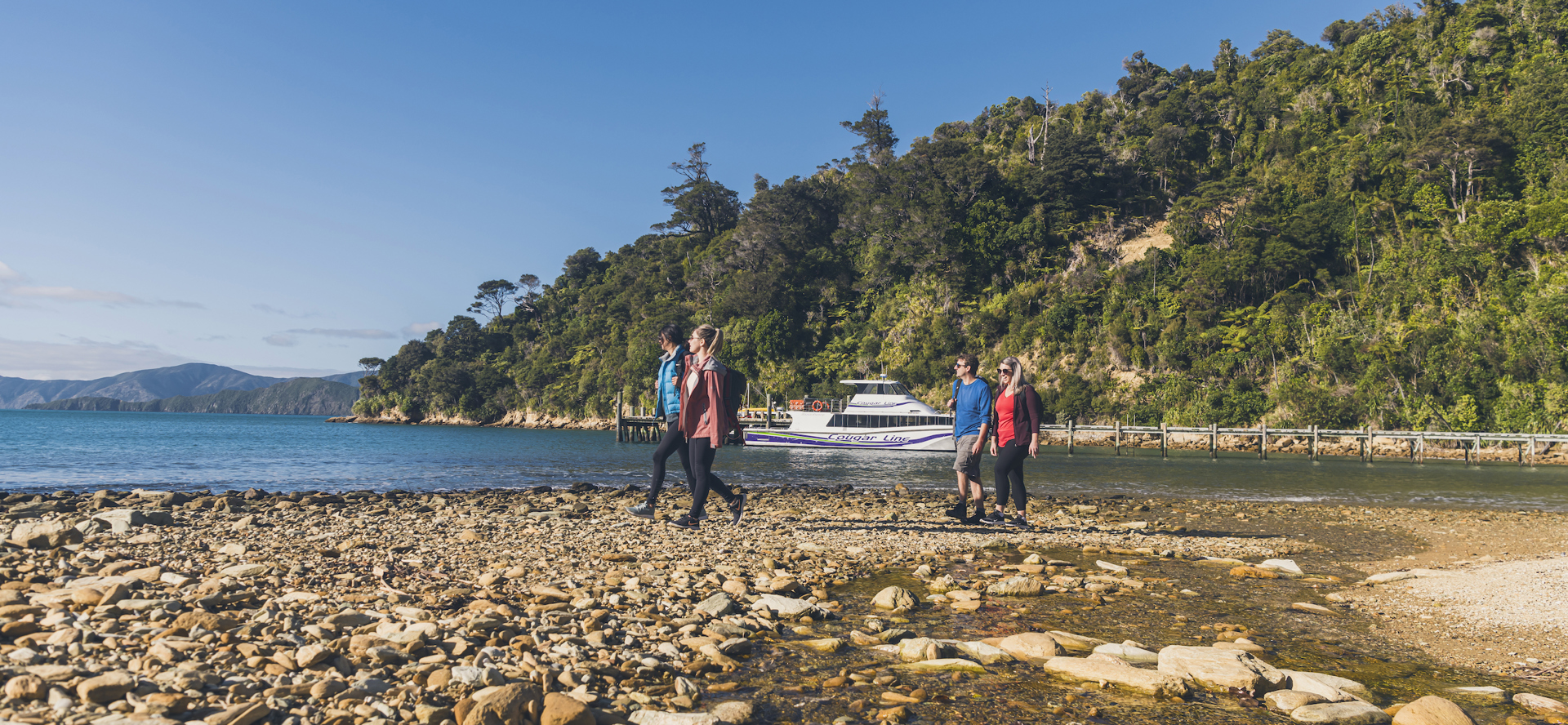 Four people walking on the beach at Ship Cove/Meretoto with a Cougar Line boat in the Marlborough Sounds, New Zealand.