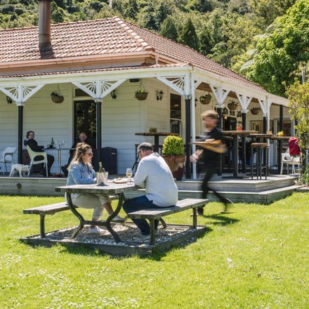 A couple sitting at the lawn picnic table receives food from a waiter in front of the Howden Homestead at the Furneaux Lodge Restaurant in the Marlborough Sounds at the top of New Zealand's South Island 