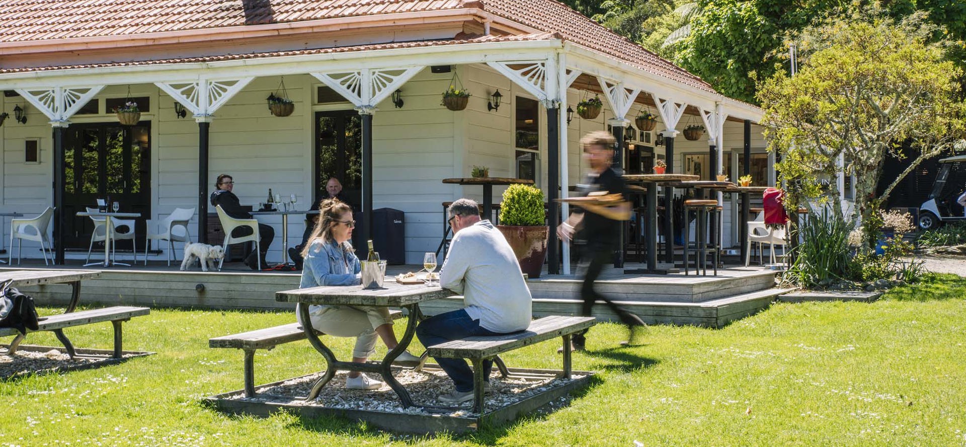 A couple sitting at the lawn picnic table receives food from a waiter in front of the Howden Homestead at the Furneaux Lodge Restaurant in the Marlborough Sounds at the top of New Zealand's South Island 