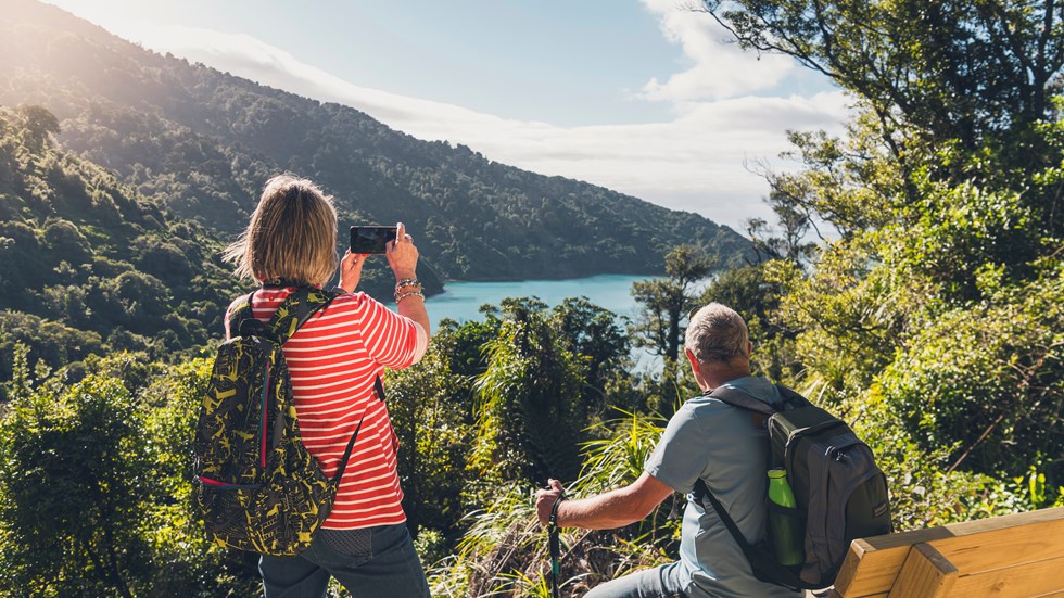 A woman takes a photo on her phone of the view across Ship Cove/Meretoto while a man sits on a bench on the northern Queen Charlotte Track in the Marlborough Sounds, New Zealand.