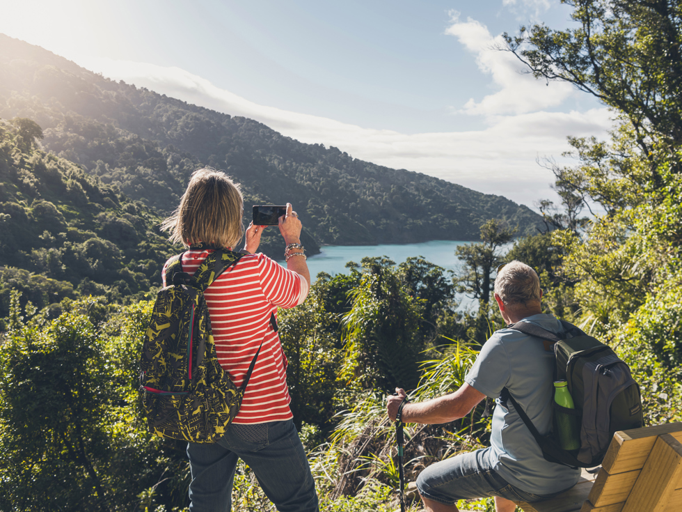 A woman takes a photo on her phone of the view across Ship Cove/Meretoto while a man sits on a bench on the northern Queen Charlotte Track in the Marlborough Sounds, New Zealand.