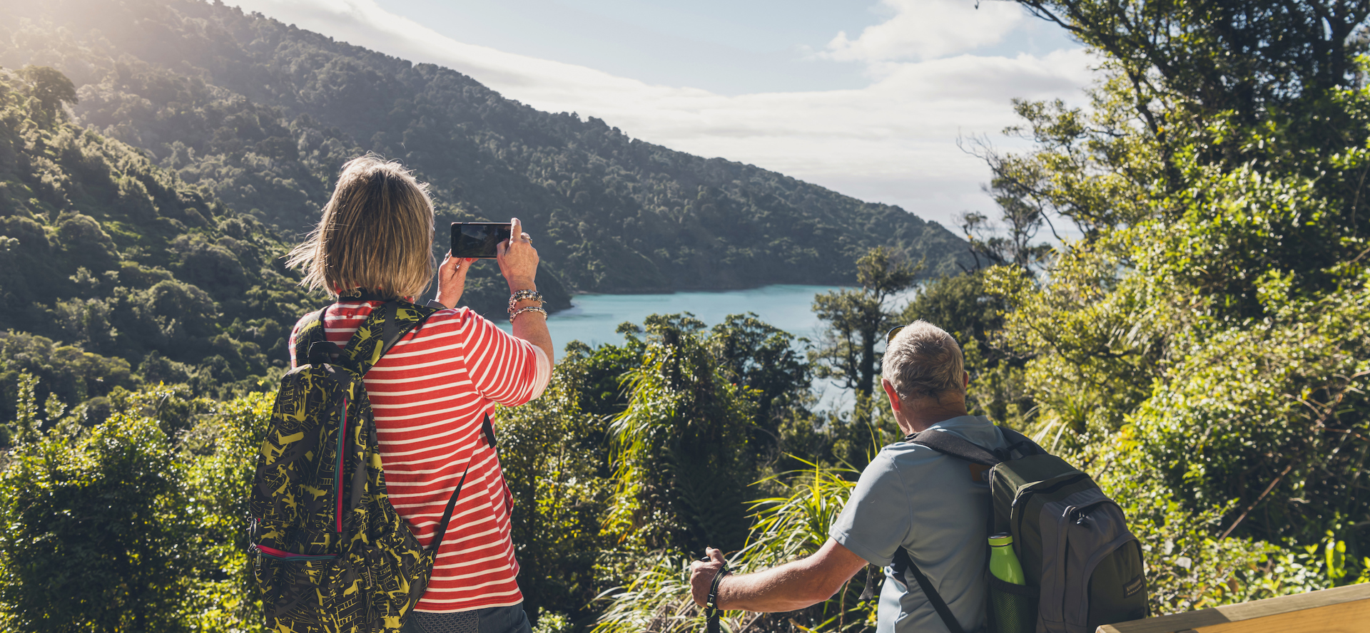 A woman takes a photo on her phone of the view across Ship Cove/Meretoto while a man sits on a bench on the northern Queen Charlotte Track in the Marlborough Sounds, New Zealand.