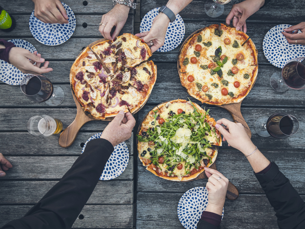 A group of friends share three hot stone baked pizzas and drinks from the Boatshed Cafe and Bar at Punga Cove located in the Marlborough Sounds of New Zealand's South Island