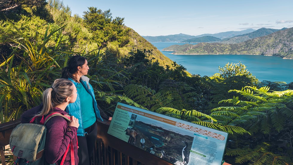 Two women admire the view over Ship Cove/Meretoto and read an interpretive sign in the outer Queen Charlotte Sound/Tōtaranui, Marlborough Sounds, New Zealand.