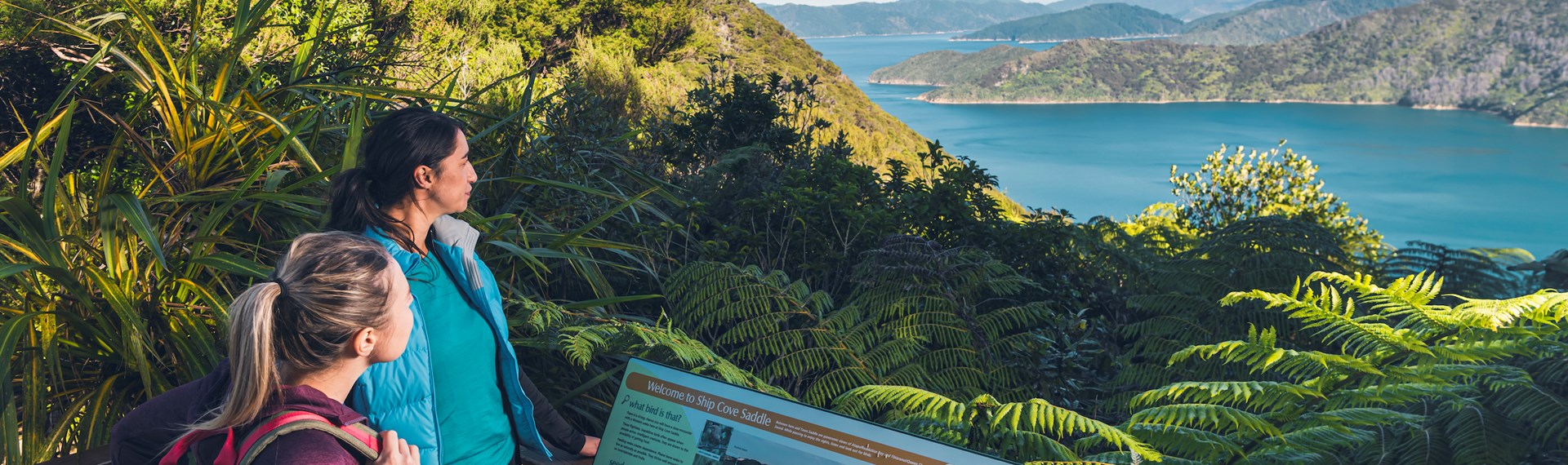 Two women admire the view over Ship Cove/Meretoto and read an interpretive sign in the outer Queen Charlotte Sound/Tōtaranui, Marlborough Sounds, New Zealand.