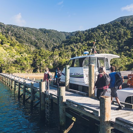 Four people walk on the jetty at Ship Cove/Meretoto next to a Cougar Line boat in the Marlborough Sounds, New Zealand.