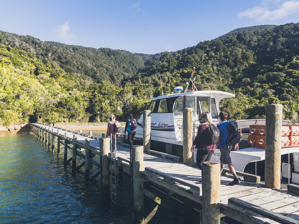 Four people walk on the jetty at Ship Cove/Meretoto next to a Cougar Line boat in the Marlborough Sounds, New Zealand.
