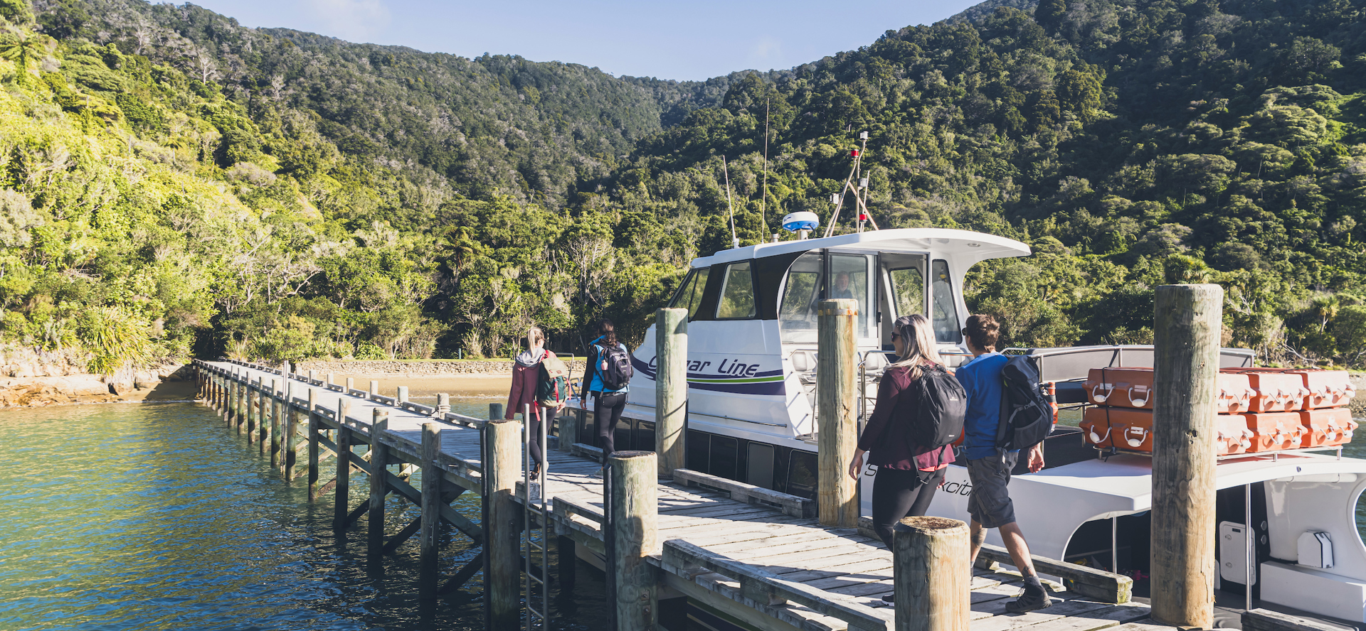 Four people walk on the jetty at Ship Cove/Meretoto next to a Cougar Line boat in the Marlborough Sounds, New Zealand.