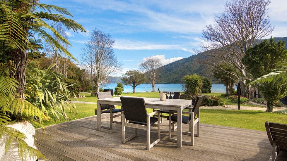 The view from a Cook's Cottage at Furneaux Lodge overlooks the furnished outdoor deck, sweeping green lawn and Endeavour Inlet in the Marlborough Sounds, New Zealand