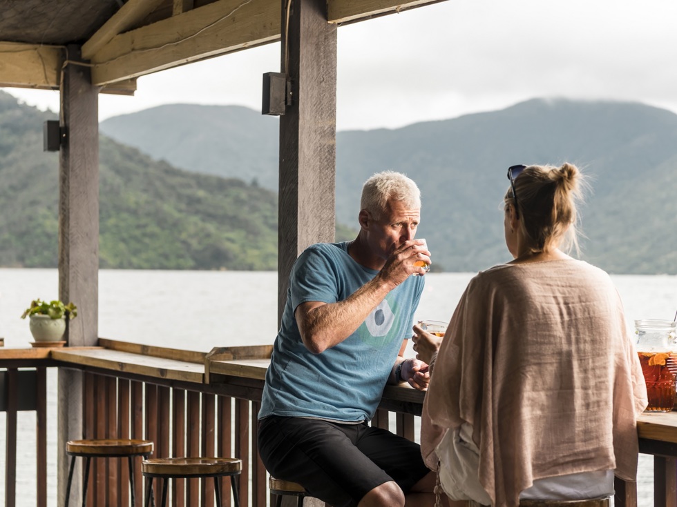 Couple relax at the Boatshed Cafe and Bar outdoor jetty bar with drinks and views of the scenic Endeavour Inlet from Punga Cove