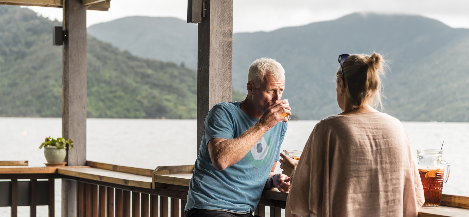 Couple relax at the Boatshed Cafe and Bar outdoor jetty bar with drinks and views of the scenic Endeavour Inlet from Punga Cove
