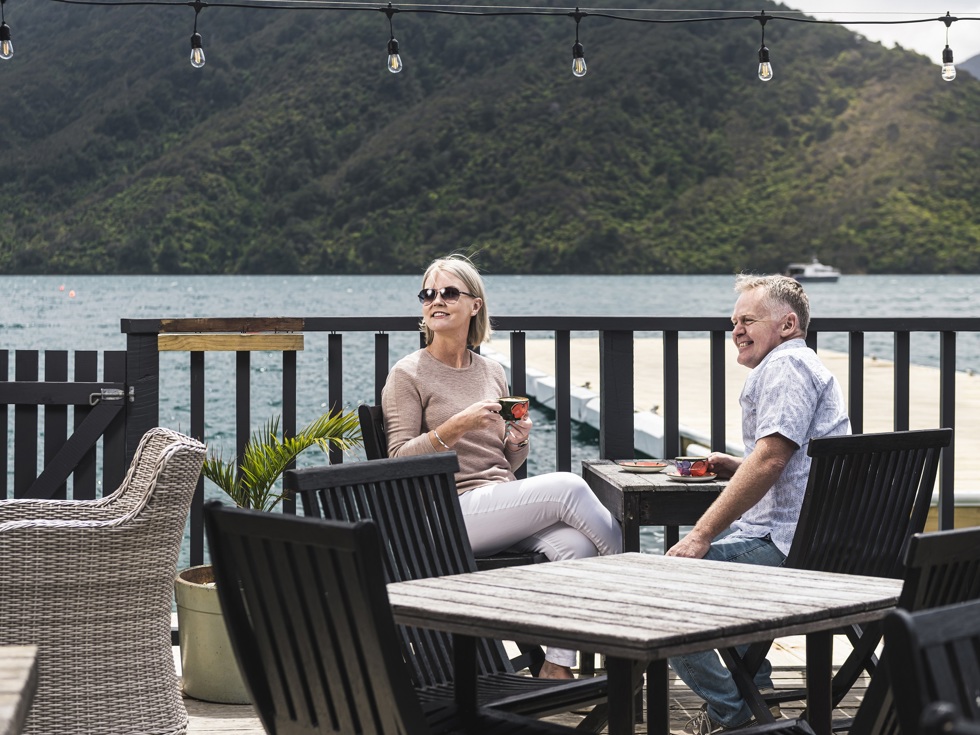 A couple sit at the Boatshed Cafe and Bar outdoor seating area with coffee enjoying the scenic views of the Endeavour Inlet in the Marlborough Sounds, New Zealand