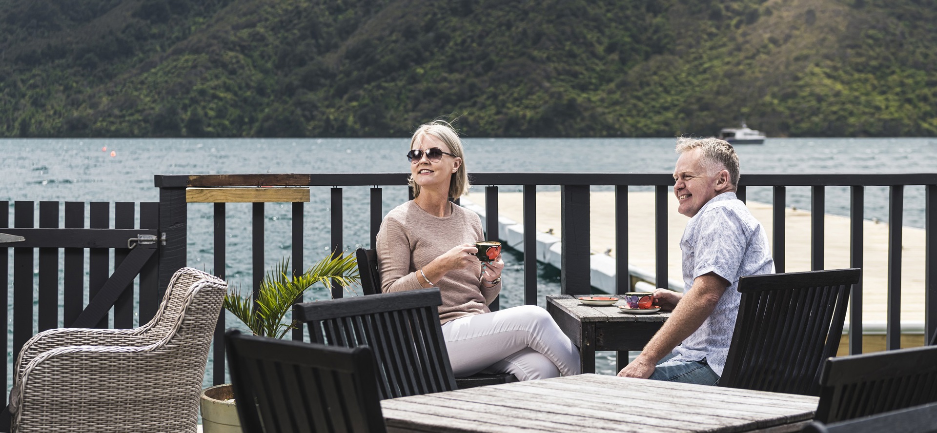 A couple sit at the Boatshed Cafe and Bar outdoor seating area with coffee enjoying the scenic views of the Endeavour Inlet in the Marlborough Sounds, New Zealand