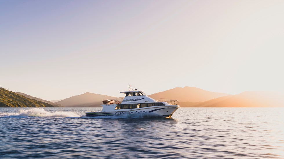 A Cougar Line boat cruises through the Marlborough Sounds under an orange sky before sunset, New Zealand.