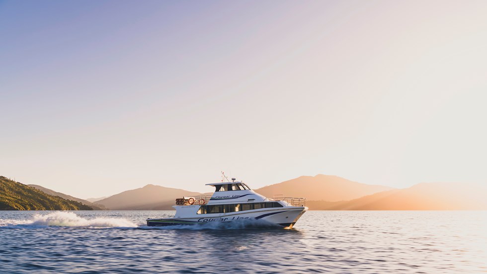 A Cougar Line boat cruises through the Marlborough Sounds under an orange sky before sunset, New Zealand.