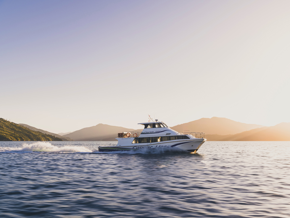 A Cougar Line boat cruises through the Marlborough Sounds under an orange sky before sunset, New Zealand.