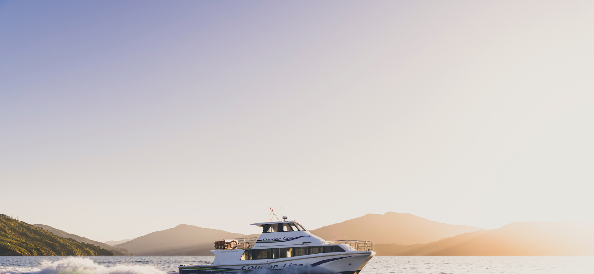 A Cougar Line boat cruises through the Marlborough Sounds under an orange sky before sunset, New Zealand.